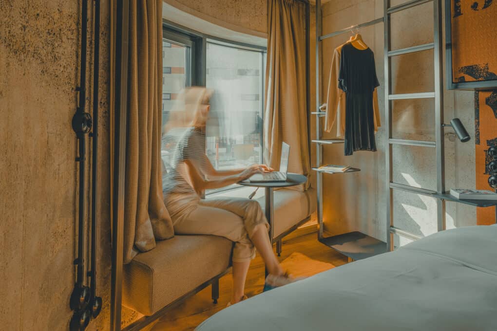 Woman sitting on the windowsill in the hotel bedroom, working on a laptop with a view outside – peaceful and bright workspace.