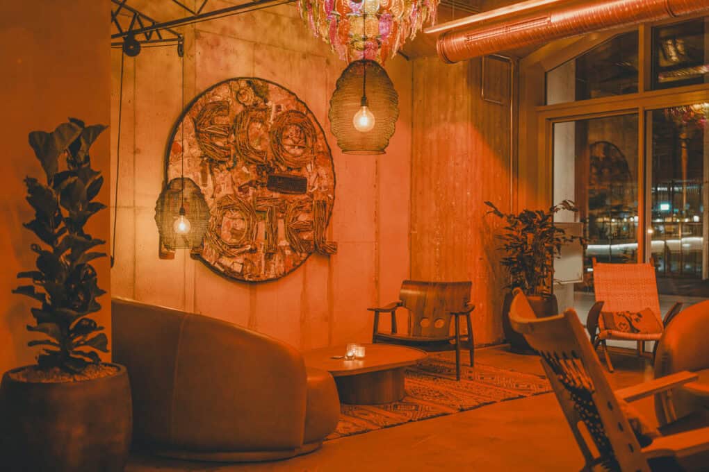 Evening mood with reddish, soft light in the John & Will lobby. View of a seating area with armchairs and an artwork made of recycled materials with the inscription “Eco Hotel.”