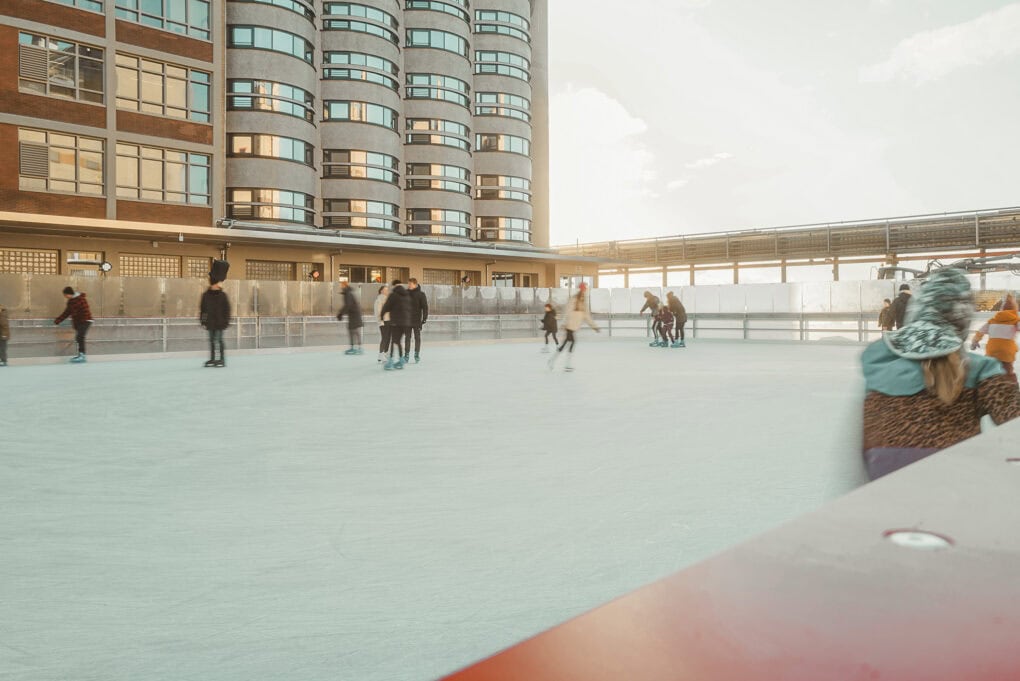 People ice skating on the rink. In the background, the John & Will can be seen.