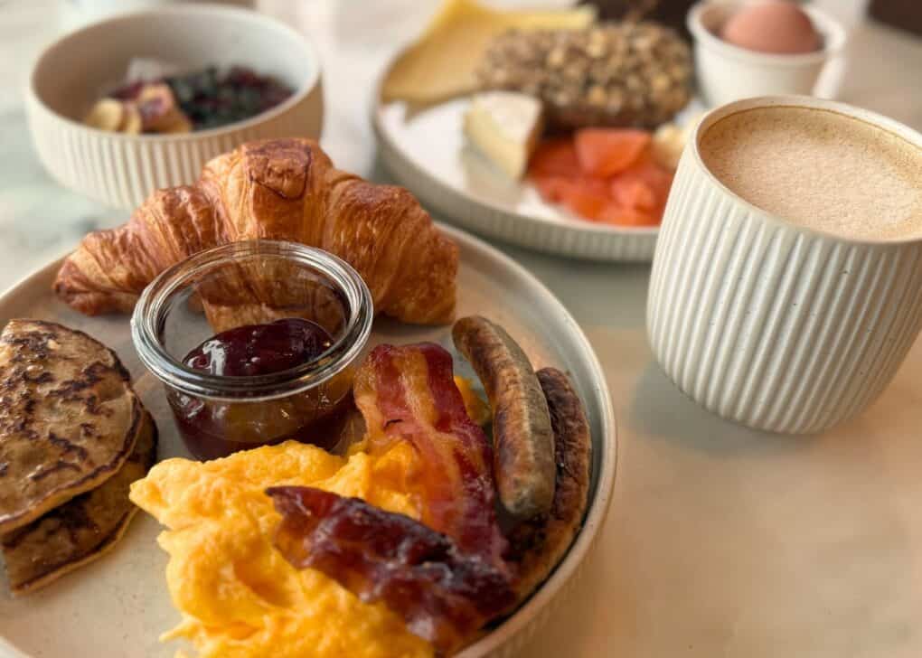 Breakfast plate with croissant, scrambled eggs, and bacon on a light marble table in the warm morning light.