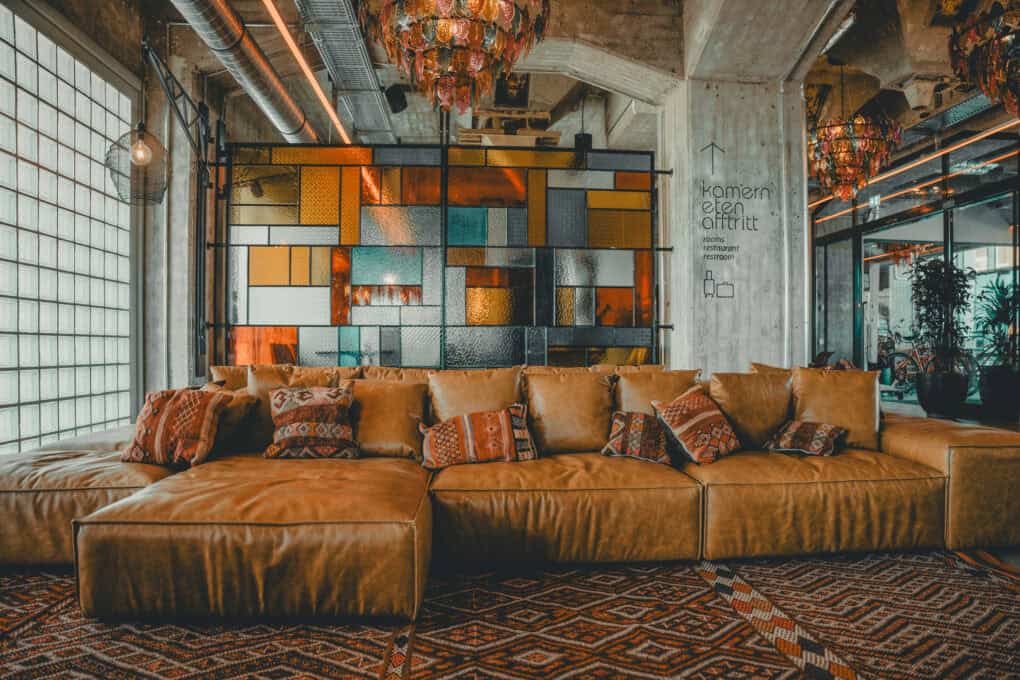 Großes gelbes Ledersofa vor buntem Glas im Barbereich der Barception im John & Will Hotel Bremen – warme, einladende Atmosphäre mit viel Sonnenlicht.
