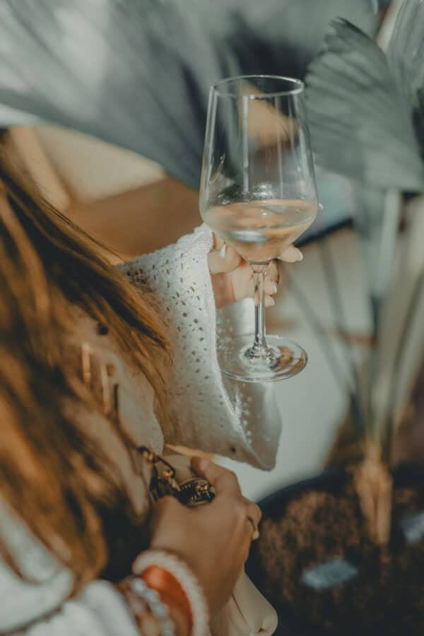 Nahaufnahme einer Frau mit langen braunen Haaren und weißem Strickpullover, die ein Glas Weißwein in der Hand hält. Das Bild vermittelt eine entspannte Atmosphäre in einem modernen Restaurant oder Hotel, ideal für Genießer und Weinliebhaber.