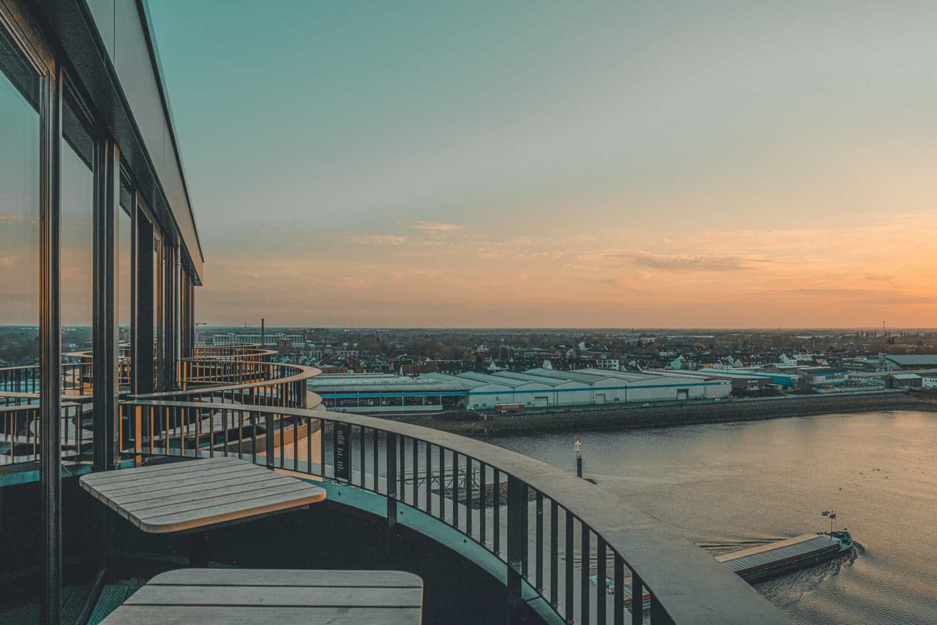Sonnenuntergang auf der Dachterrasse des John & Will Hotels Bremen mit Blick über die Weser – stilvolle Sitzplätze entlang der Glasfassade und eindrucksvolle Aussicht auf den Hafen und das Umland. Perfekte Kulisse für Sundowner und entspannte Momente über den Dächern der Überseestadt.