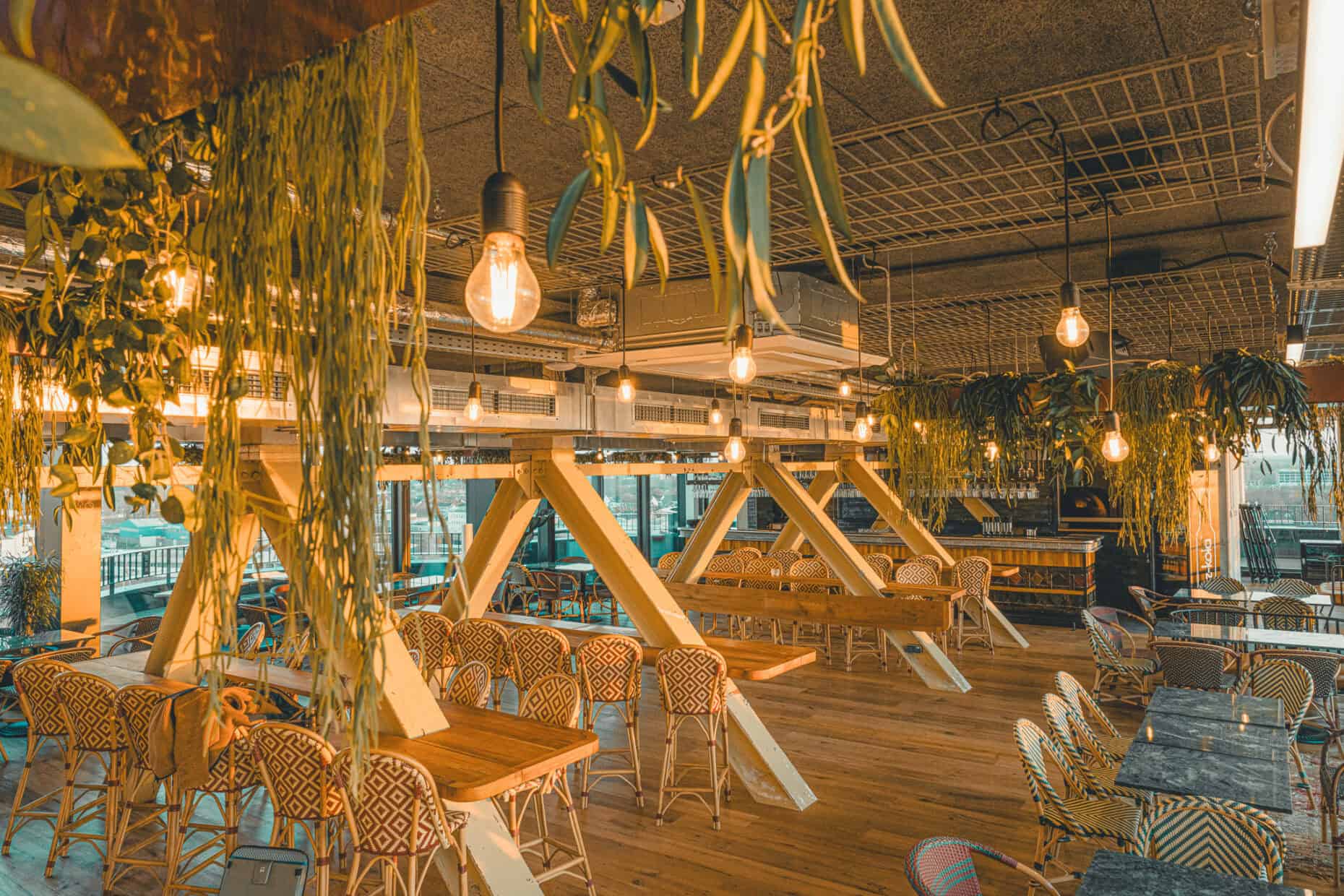 Stilvolles Rooftop-Restaurant im John & Will Hotel in Bremen mit hängenden Pflanzen, Vintage-Glühbirnen, Holztischen und gemusterten Rattanstühlen – Industrial-Design trifft auf urbane Gemütlichkeit mit Ausblick über die Weser.