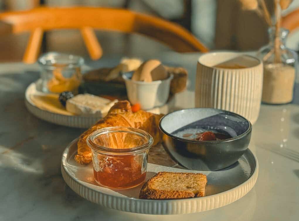 Frühstücksteller mit Croissant, Marmelade, Joghurt mit Beeren, Toast, Käse und einem gekochten Ei auf einem hellen Marmortisch im warmen Morgenlicht.
