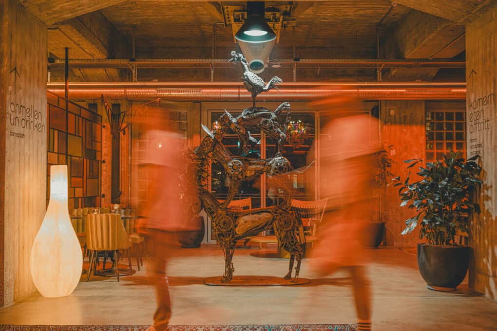 Kunst in der Lobby des John & Will: Bei leicht rötlichem Licht sieht man die Bremer Stadtmusikanten-Skulptur aus Schrott. Davor bewegen sich zwei Personen in der Unschärfe.